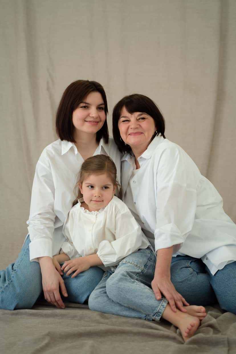 Three generations of women in matching white shirts posing together for a family portrait: a grandmother, her adult daughter, and her young granddaughter